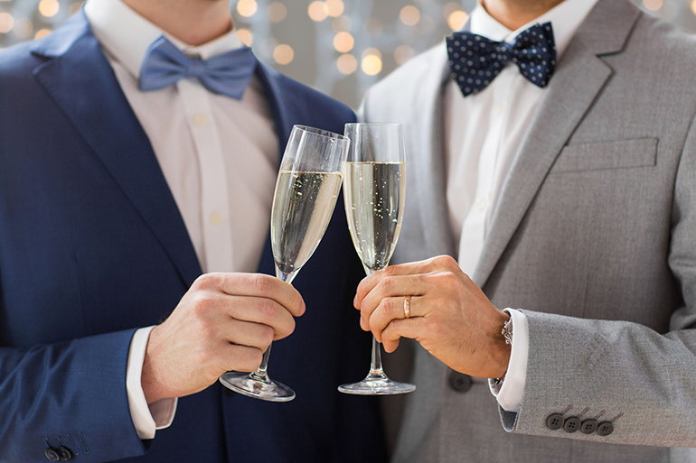 people, celebration, homosexuality, same-sex marriage and love concept - close up of happy married male gay couple drinking sparkling wine on wedding over holidays lights background