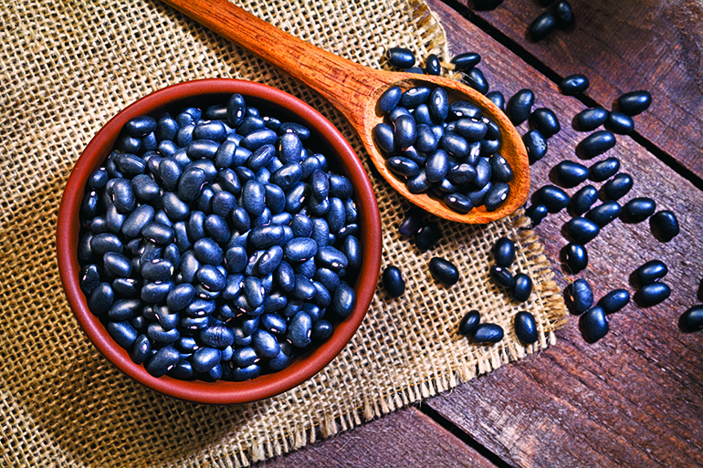 Top view of a brown bowl filled with black beans shot on rustic wood table. A wooden spoon is beside the bowl with some beans on it. DSRL studio photo taken with Canon EOS 5D Mk II and Canon EF 100mm f/2.8L Macro IS USM