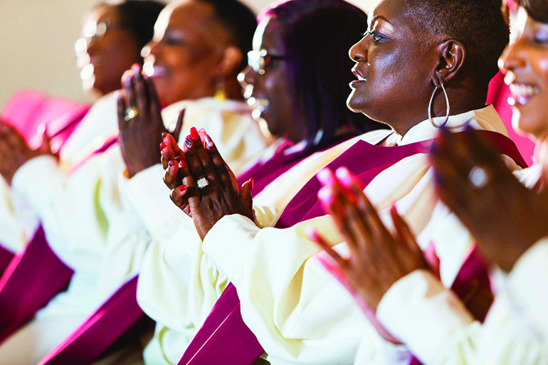 A group of mature black women in church robes, sitting in a row, clapping. They are members of the church choir listening to a sermon.