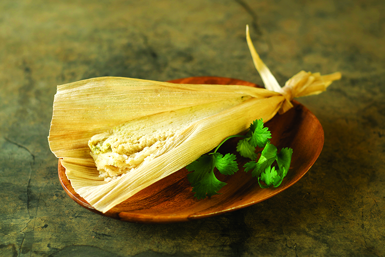 Mexican chicken tamale in corn husk on wood plate garnished with cilantro.