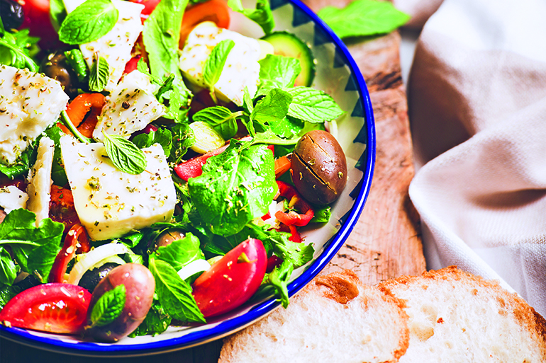 Traditional Greek salad with feta cheese, olive oil and olives and glass of red wine over rustic wooden serving board on white marble table, selective focus. Mediterranean local cuisine