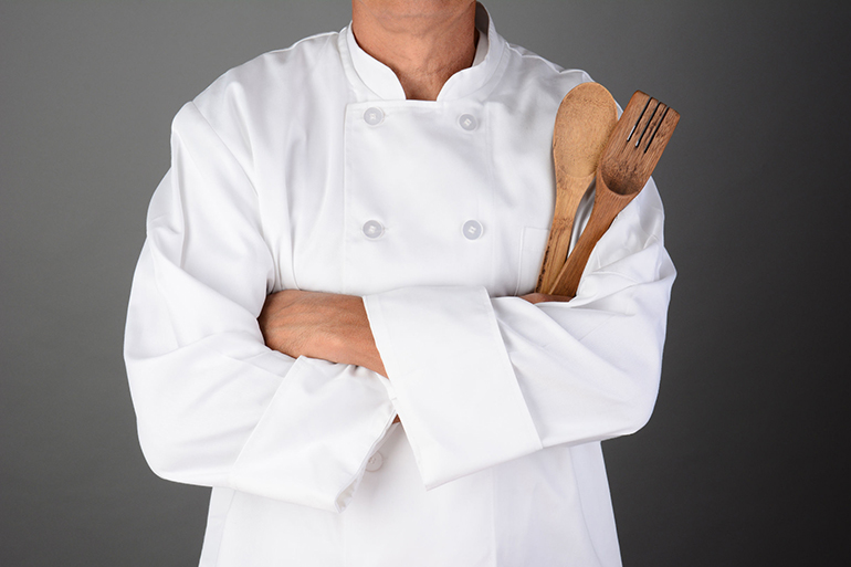 closeup of a chef with his arms folded holding wood utensils man is unrecognizable horizontal format on a light to dark gray background model released