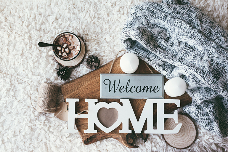 Winter homely scene, scandinavian style. Warm knit sweater, candles, cup of sweet cocoa with marshmallows and other decor on tray in bed. Wooden craft letters Welcome Home. Lazy cold weekend.