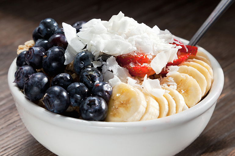Blueberries, bananas, and coconut on top of acai served in a white bowl.