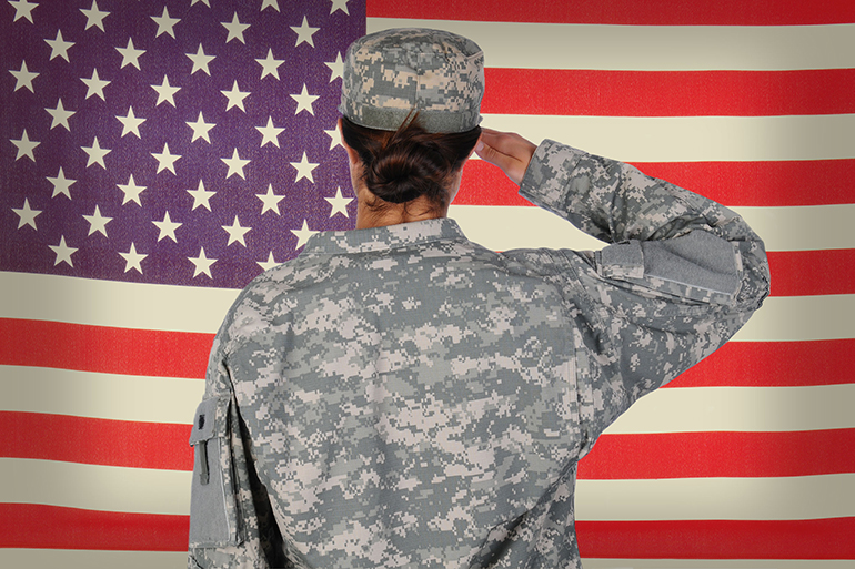 Female Soldier standing in front of and saluting an American flag. Woman is seen form behind only showing her from the waist up. Horizontal format.