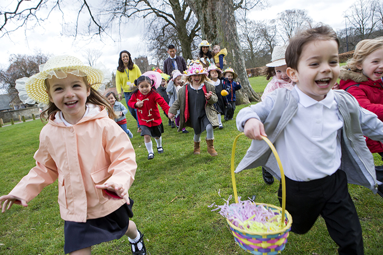 Nursery children running across a field during their outdoor Easter egg hunt, they are wearing handmade hats and carrying baskets.