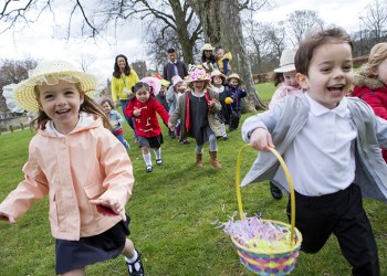 Nursery children running across a field during their outdoor Easter egg hunt, they are wearing handmade hats and carrying baskets.