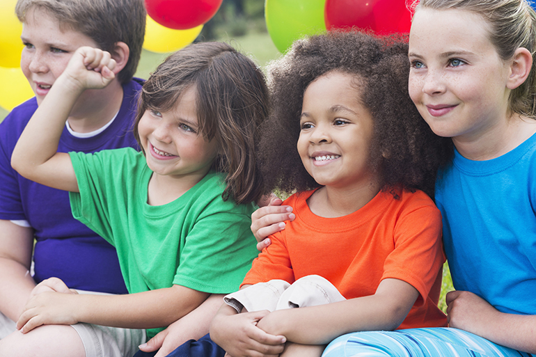 A multi-ethnic group of children, ages 5 to 12, in the park, having fun in summer camp. They are wearing colorful shirts, sitting together, watching something. Colorful balloons are behind them.