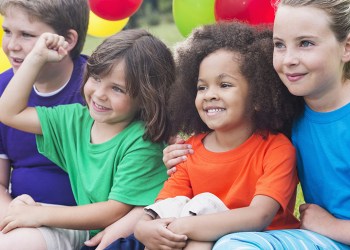 A multi-ethnic group of children, ages 5 to 12, in the park, having fun in summer camp. They are wearing colorful shirts, sitting together, watching something. Colorful balloons are behind them.