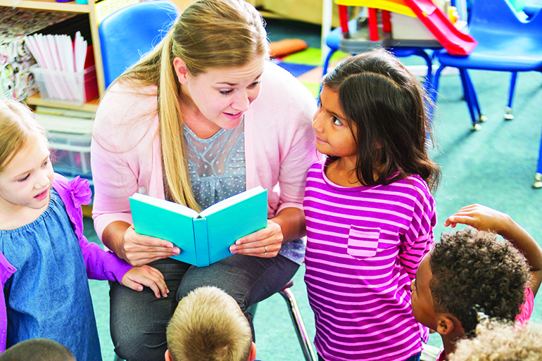 A preschooler teacher in the classroom reading to her class. The multiracial children are sitting on the floor looking up at her. She is talking to a little girl of Pacific Islander ethnicity who is kneeling beside her.