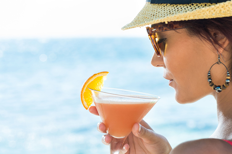 Closeup Of Young Woman Wearing Hat And Sunglasses Holding Cocktail Glass At Seaside
