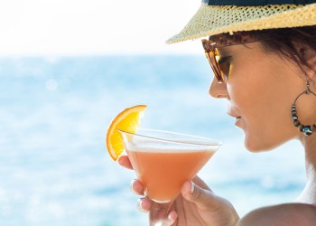 Closeup Of Young Woman Wearing Hat And Sunglasses Holding Cocktail Glass At Seaside