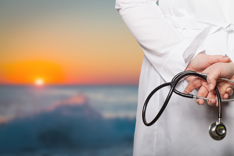 The doctor put his hands with stethoscope behind his back on a blurred background.