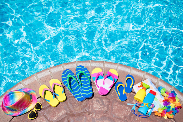 Swimming pool accessories flat lay. Top view of beach items on pool deck. Flip flops, bikini and hat, sun glasses. Water toys. Summer vacation in tropical resort. Copy space. Colorful beach wear.