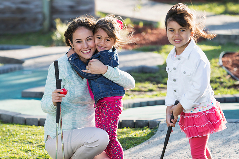 A young family playing miniature golf outdoors on a sunny day. A Hispanic mother with her two daughters, 3 and 4 years old. She is hugging the younger girl, while the older one is swinging a golf club.