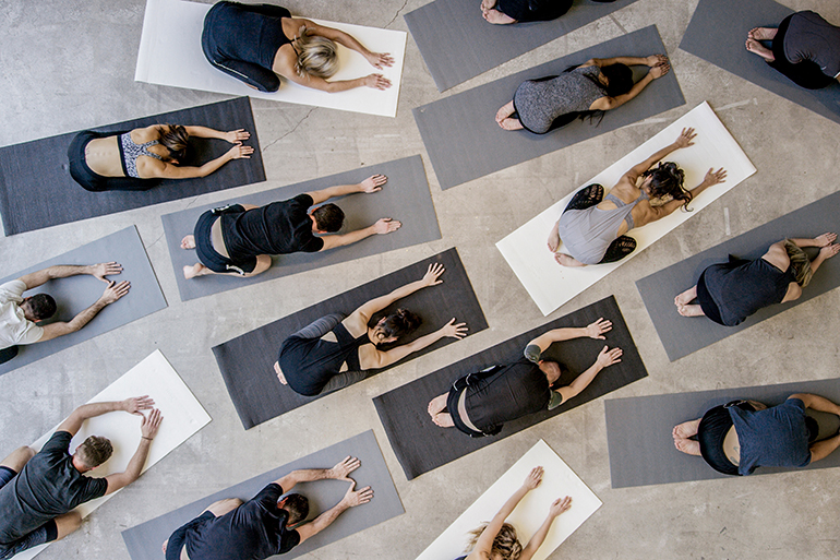 An aerial shot of a multi ethnic group of men and women practice yoga on on mats while wearing grey, black and white in an industrial setting. They are reaching forward in child's pose.