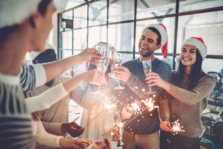 Merry Christmas and Happy New Year 2018!Multiracial young creative people are celebrating holiday in modern office. Group of young business people are drinking champagne with sparkling bengal lights in coworking. Successful hipster team of freelancers.