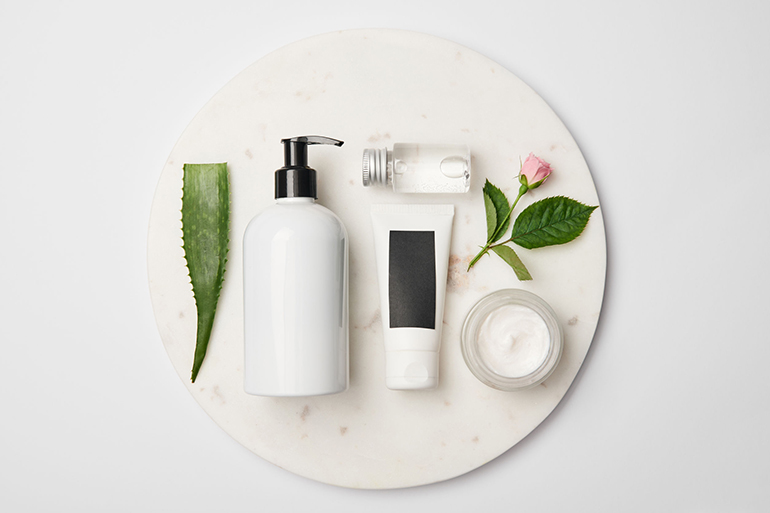 Top view of different cosmetic containers, aloe vera leaf and rose flower on white round surface