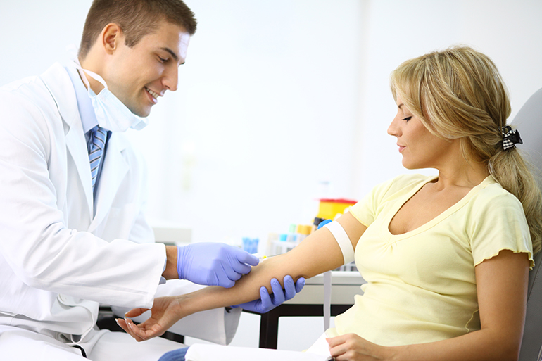 Middle aged blond woman visiting general practitioner.Both are sitting in examination room.The doctor is drawing blood sample from her vein.She has dark blond hair pulled back with a clip.There's a little smir on her face because this is painless procedure.