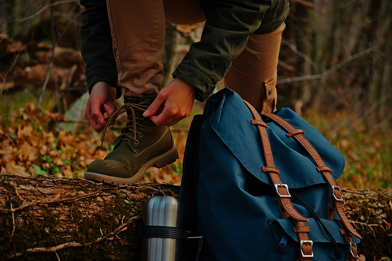 Female hiker tying shoelaces outdoors in autumn forest, near thermos and backpack. View of legs. Hiking and leisure theme