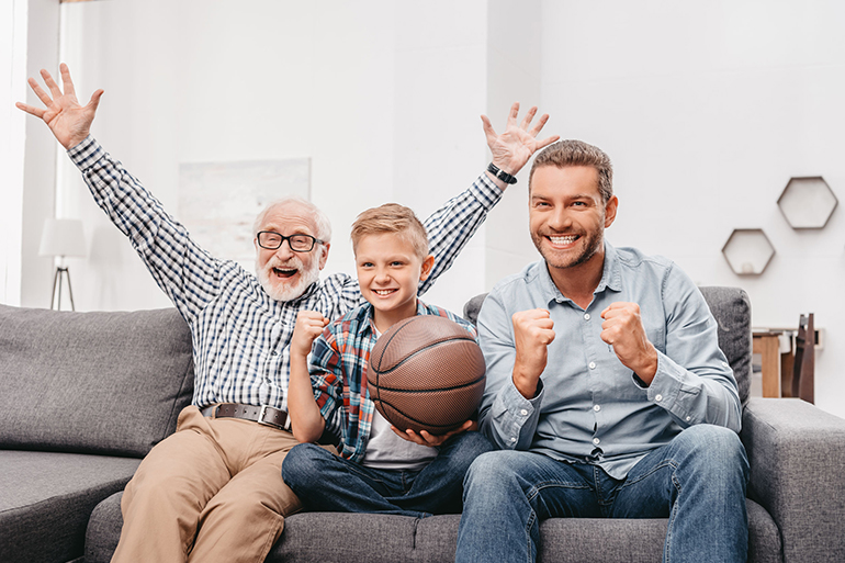 Little boy on couch with grandfather and father, cheering for a basketball game and holding a basketball ball