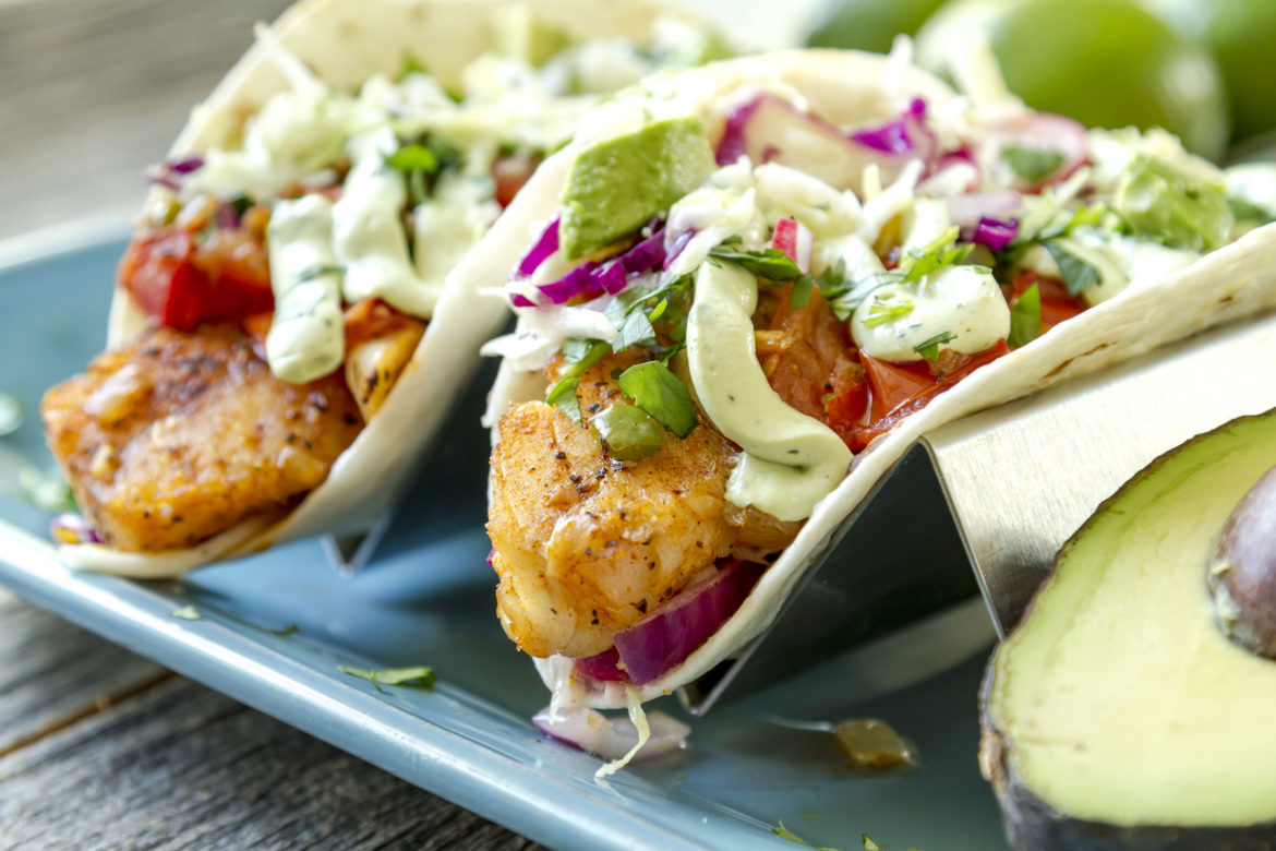 Close up of fresh fish tacos with coleslaw, avocado, salsa and lime creme in a flour tortilla on blue plate