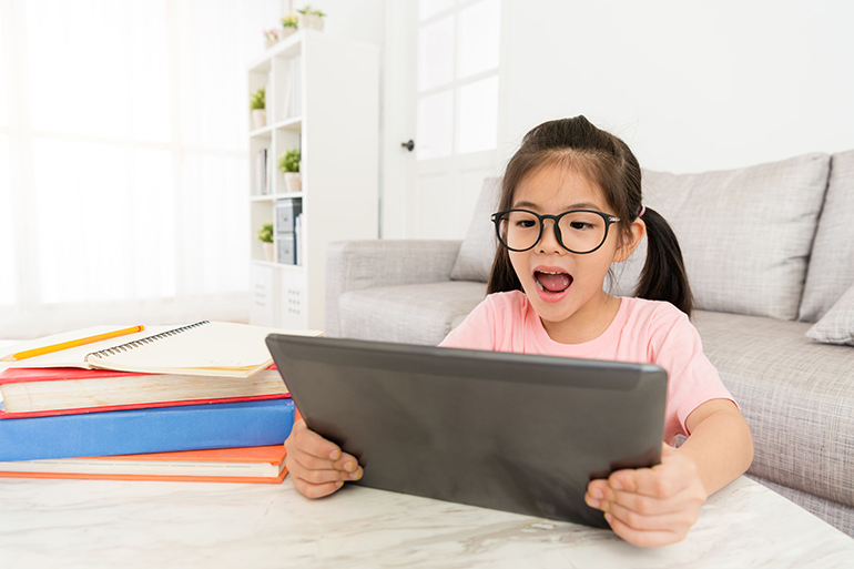 cheerful young female children holding mobile digital tablet sitting in living room and using online e-learning system studying.