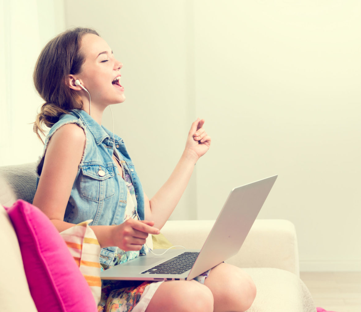 Beautiful young woman sitting on sofa at home, using laptop computer, laughing and having fun. Happy young woman enjoying the music with headphones and dancing. Emotions. Happiness. Good mood