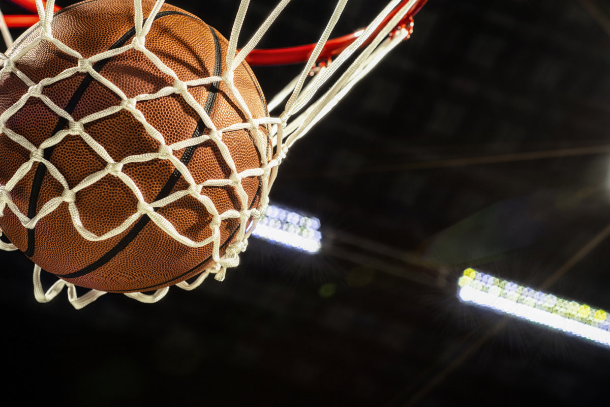 A close-up view looking up at an orange basketball falling through the rim and a white nylon net with the arena lights and lens flare in the background.