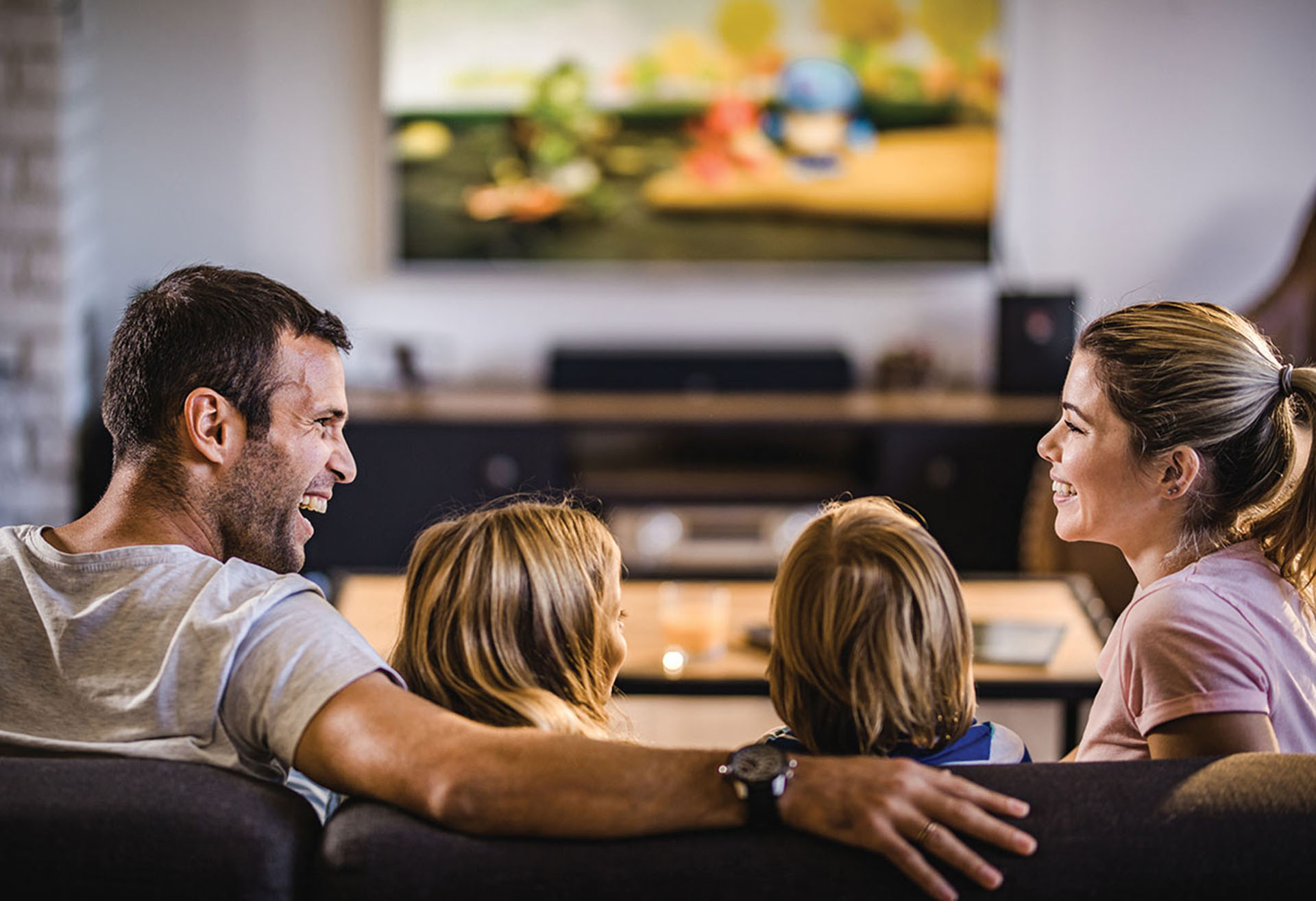 Happy parents talking while watching TV with their kids at home.