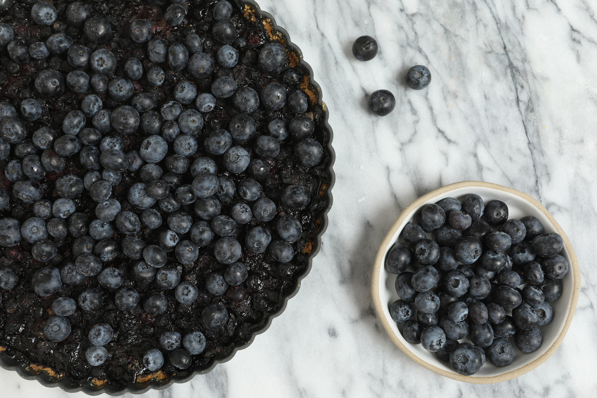 An overhead extreme close up horizontal photograph of a homebred double blueberry tart and a small white bowl of fresh blueberries.