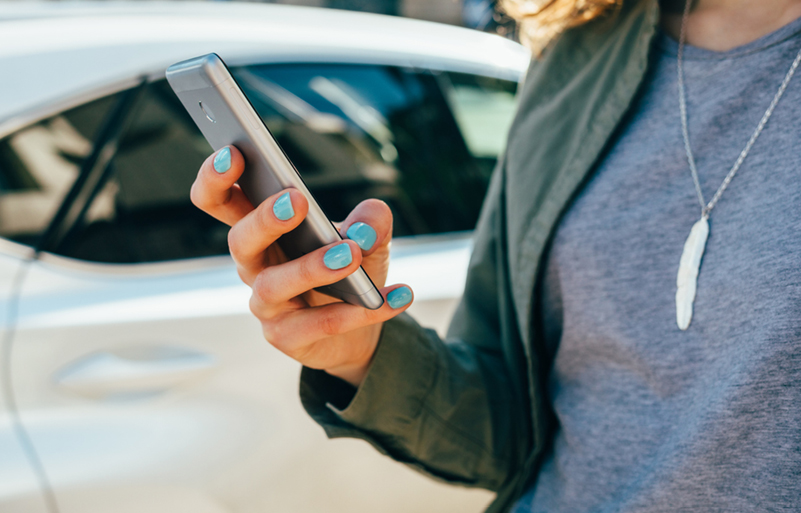 Young woman using smart phone standing on street near road and car. Female's hands holding mobile device in city, close-up.