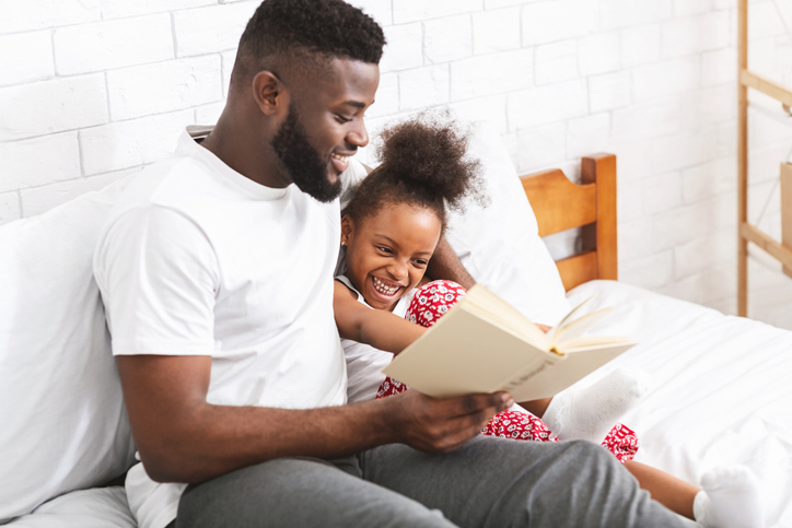 Story time. Loving african american dad reading fairy tales to his little daughter, sitting together on bed