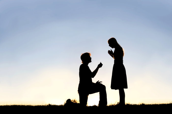 A silhouette of a young man, down on one knee and holding a diamond engagement ring, proposing to his girlfriend for engagement and wedding
