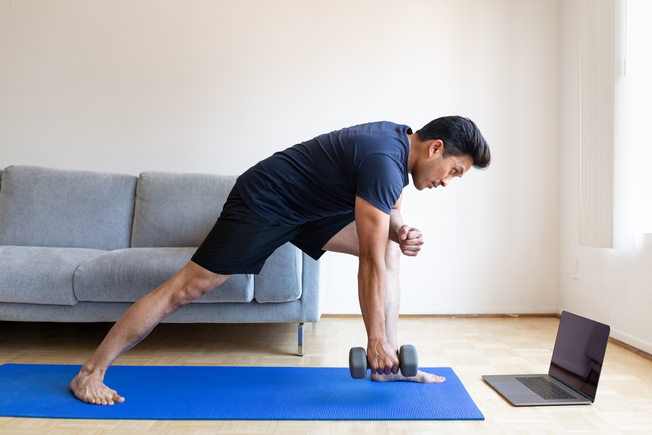 Man watching video tutorial on laptop and doing dumbbell workout at home. Man exercising with dumbbell and watching online fitness live streaming classes on his laptop.