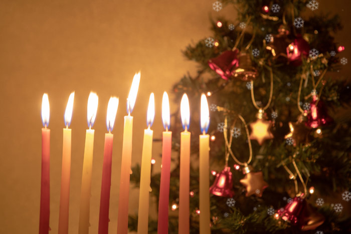 Hanukkah candles burning on the background of Christmas tree, decorated with bells and stars of David. Concept of two holidays happening in the same period this year. Be the Light