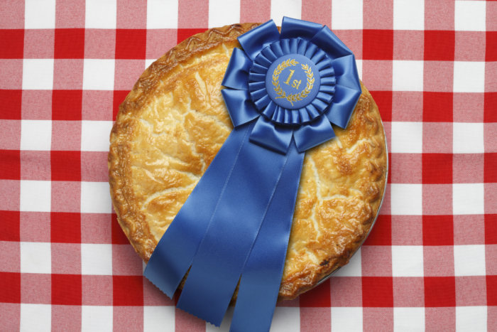 Best of the Best pie with a blue first place ribbon on top. The pie is sitting on a classic red checkerboard tablecloth. Image evokes the feeling of classic Americana. Camera angle is from directly above.
