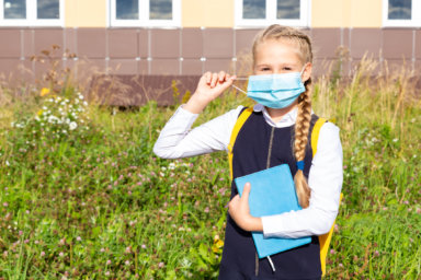 A girl in a blue school uniform stands in front of the school building in a medical mask, holds a textbook in her hands, looks at the camera. Back to school. Security concept, virus protection, new normal.
