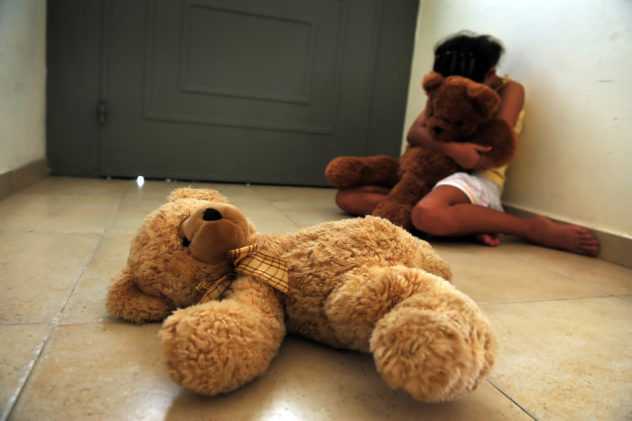A young girl who is a victim of domestic violence and sexual abuse sits on the floor next to the front door and gets comfort from her teddy bears