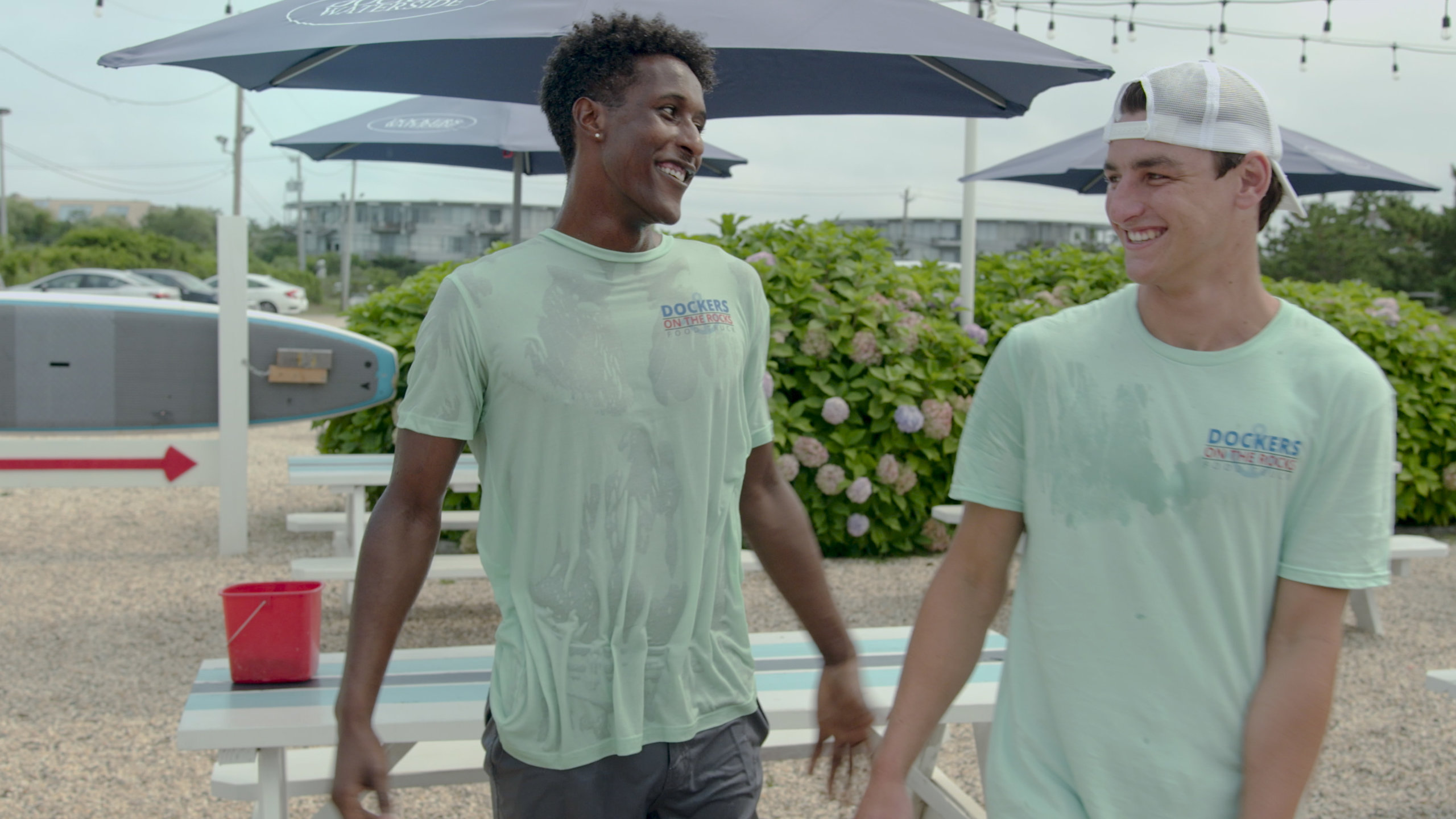 Scene from Forever Summer: Hamptons guys wearing Dockers on the Rocks food truck shirts