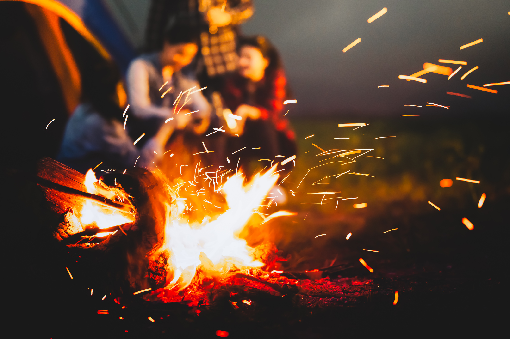 Sparking bonfire with tourist people sit around bright bonfire near camping tent in forest in summer night background. Group of student at outdoor fire fuel. Travel activity and long vacation weekend