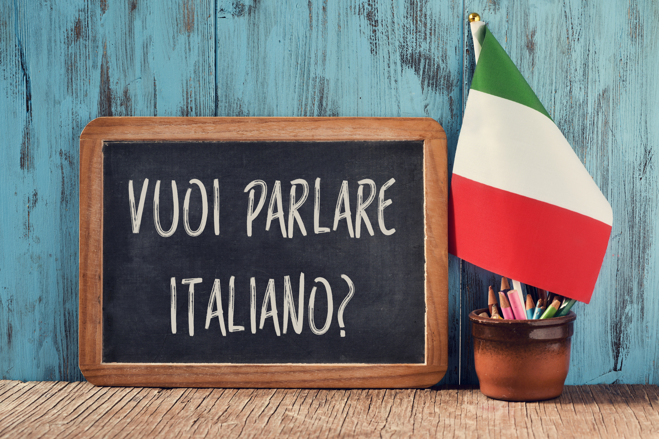 a chalkboard with the question vuoi parlare italiano?, do you want to speak Italian? written in Italian, a pot with pencils, some books and the flag of Italy on a wooden desk