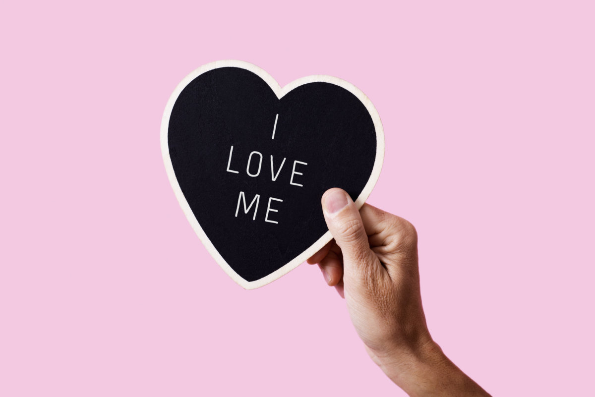 closeup of a man holding a black heart-shaped sign with the text I love me written in it, on a pink background -love yourself