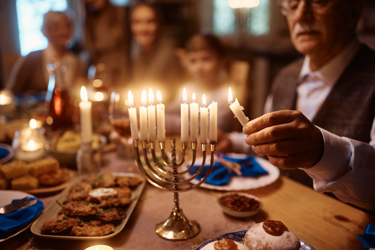 Close up of extended Jewish family celebrating Hanukkah Chanukah at dining table. Focus is on mature man lighting candles in menorah.