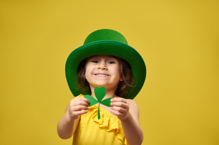Pretty little girl holds a clover leaf in hands and shows it to camera. Saint Patrick's Day concept for kids on yellow background.