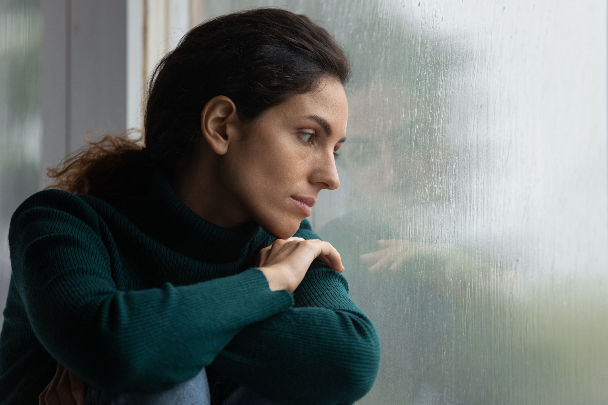 Thoughtful stressed young hispanic latin woman sitting on windowsill, looking outside on rainy weather, having depressive or melancholic mood, suffering from negative thoughts alone at home. loneliness