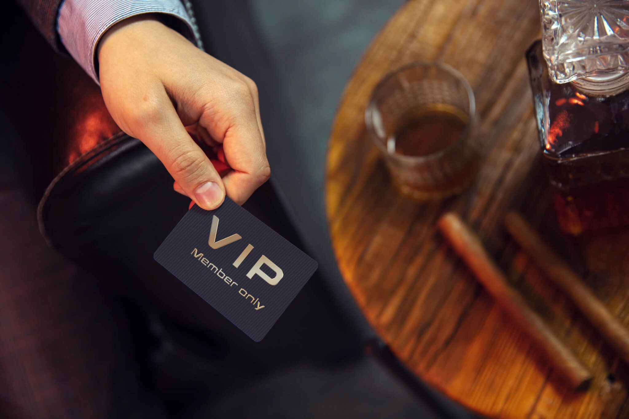 Man holds VIP member card. View from the top on the gentleman's hand that holds exclusive VIP membership card next to the wooden table with whisky in carafe and glass with cigars.