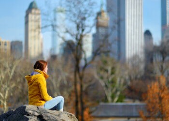 Beautiful young woman looking at skyscrapers while sitting on a rock in Central Park, New York