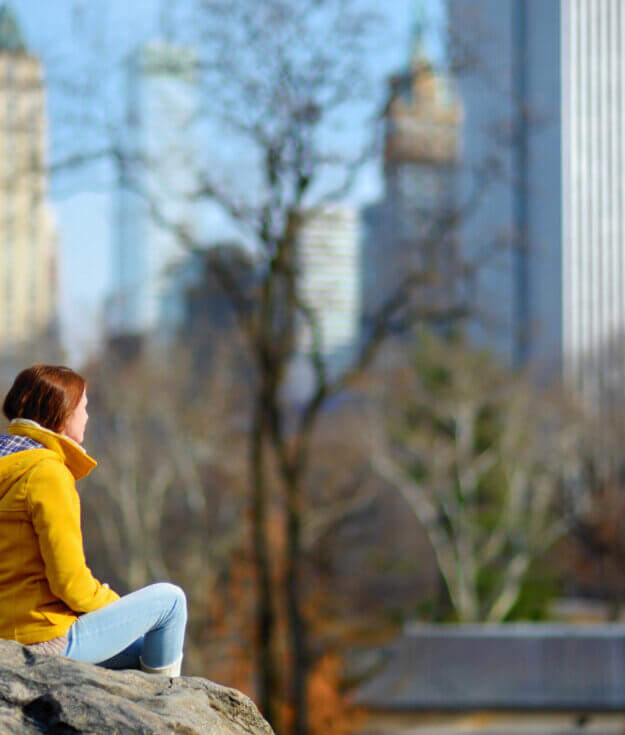Beautiful young woman looking at skyscrapers while sitting on a rock in Central Park, New York