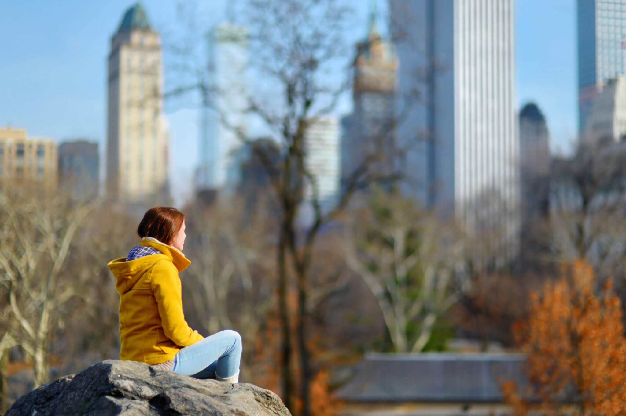 Beautiful young woman looking at skyscrapers while sitting on a rock in Central Park, New York